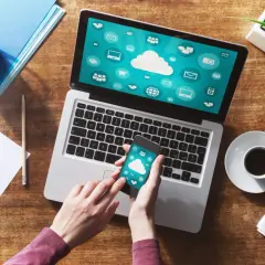 Dłonie trzymające smartfon z ikonami chmury na tle laptopa z podobnymi ikonami.