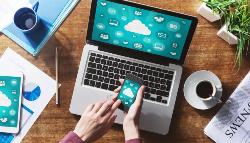Dłonie trzymające smartfon z ikonami chmury na tle laptopa z podobnymi ikonami.